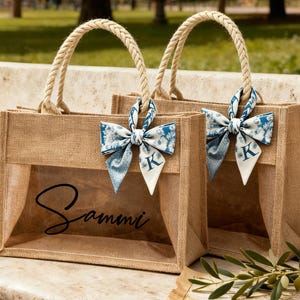 Op de afbeelding: Twee jute draagtassen met transparante voorpanelen. Elke tas heeft een gevlochten touwhandvat en een blauw-witte bloemenstrik met een wit label met de letter "K". Eén tas heeft de naam "Sammi" in het zwart gedrukt.