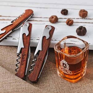 May include: Three wooden bottle openers with engraved names. The openers have a corkscrew, bottle opener, and a small knife. A small glass of amber liquid sits on a burlap surface.
