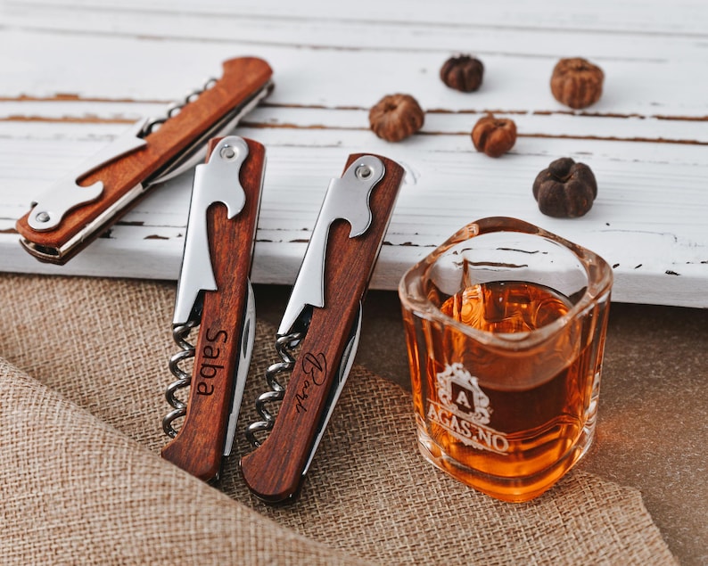 May include: Three wooden bottle openers with engraved names. The openers have a corkscrew, bottle opener, and a small knife. A small glass of amber liquid sits on a burlap surface.
