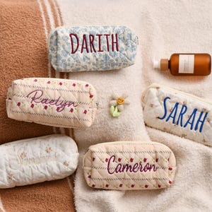 May include: Five quilted cosmetic bags with embroidered names in different colours. The bags have floral patterns in white, cream, and blue. A brown bottle with a pump and a flower-shaped pin are also visible.