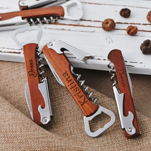 May include: Three personalized wooden bottle openers with corkscrews. The bottle openers are engraved with the names "Donna", "Kristine", and "Denise".