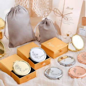 May include: An assortment of personalized compact mirrors in gold, silver, and rose gold, each engraved with a name and floral design. They are displayed with matching gift boxes and drawstring pouches, ideal for gifts.