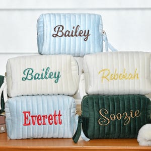 May include: Several personalised cosmetic bags in shades of light blue, white, and dark green. Each bag has a name embroidered on it in a contrasting colour. They have a ribbed texture and a zip closure.