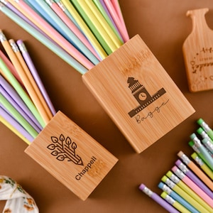 Peut inclure: Trois porte-crayons en bois avec des motifs gravés. Le premier porte-crayon a un motif d'arbre et de crayon avec le nom "Chappell" gravé. Le deuxième porte-crayon a un motif de tour d'horloge avec le nom "Bagge" gravé. Le troisième porte-crayon a un motif floral avec le mot "Amazing" gravé.