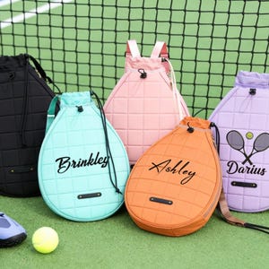 May include: Tennis-racket-shaped backpacks in black, teal, pink, orange, and lavender, with personalized names and tennis-themed graphics. A yellow tennis ball and blue tennis shoes are also visible.