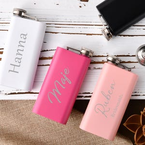 May include: Three personalised stainless steel hip flasks. One is white with the name "Hanna" engraved on it, one is pink with the name "Maie" engraved on it, and one is light pink with the name "Ruben Bridesmaids" engraved on it.