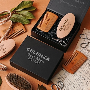 May include: A collection of grooming products, including wooden combs, a brush, and an oval-shaped item, arranged on a brown surface. The items are in black boxes with the text "CELENZA Best Man 06.12.25". Some combs are engraved with names. Scissors and greenery are also present.