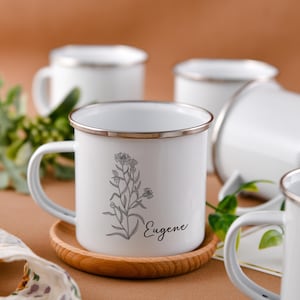 Peut inclure: Une tasse en émail blanc avec un bord argenté et une illustration en noir et blanc d'une fleur avec le nom "Eugène" imprimé sur le côté. La tasse est posée sur un dessous de verre en bois.