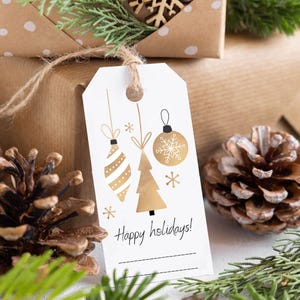 Op de afbeelding: Een wit cadeaulabel met gouden kerstversieringen en de tekst "Happy holidays!". Het label is met touw aan een bruin verpakt cadeau bevestigd. Pijnappels en groenvoorziening staan op de voorgrond.