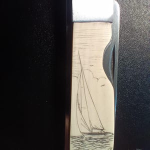May include: A folding pocket knife with a cream-colored handle featuring a detailed black ink line drawing of a sailboat on the water. The knife has a metallic bolster at the top and a partially visible blade edge along the right side of the handle.