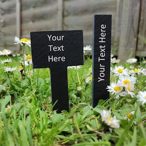 May include: Two black slate garden markers with white text reading "Your Text Here". One is rectangular, the other T-shaped. They are set in grass with white daisies. Ideal for personalised garden labels.