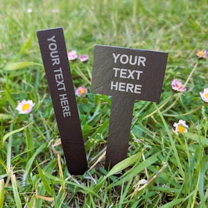 May include: Two dark grey slate garden markers with the text "YOUR TEXT HERE" in white. One marker is rectangular, and the other is a signpost shape. The markers are set in green grass with small pink and yellow flowers.