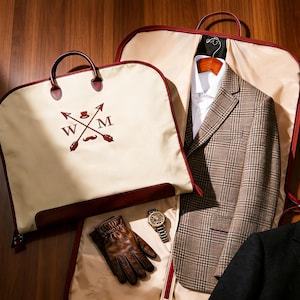 Puede incluir: Bolsa de ropa color crema con ribetes y asas marrones, con un logotipo bordado marrón con flechas, un bigote y las letras "W" y "M". Una blazer a cuadros y una chaqueta de traje negra son visibles dentro de las bolsas abiertas. Los accesorios incluyen guantes de cuero marrones y un reloj de pulsera.