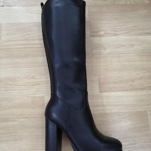 May include: A tall, black boot with a block heel and platform sole. The boot has a smooth, leather-like texture and a rounded toe. The heel measures approximately 10 cm.