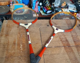 May include: Two vintage badminton rackets with wooden frames and white, red, and black accents. The rackets have a classic design with a net strung across the oval head. The handles are wrapped in black grip tape.