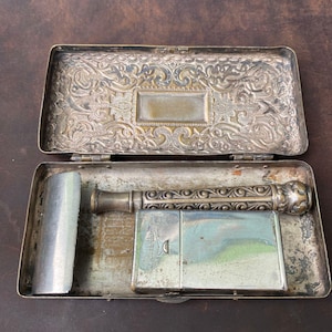 Puede incluir: Un estuche antiguo plateado abierto con tapa abisagrada, que contiene una navaja de afeitar, un pequeño espejo rectangular y un mango cilíndrico. La tapa y el mango presentan adornos de volutas. El estuche descansa sobre una superficie marrón.