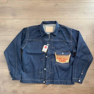 May include: A dark blue denim jacket with a button-up front, a collar, and a chest pocket. The jacket has a small tag attached and a label on the pocket that reads "SUVIN GOLD". The jacket is made of a sturdy denim material.