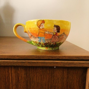 May include: Yellow ceramic teacup with a curved handle. The cup features a painted scene of two children holding hands against a sunset backdrop. The base of the cup has a green floral design.
