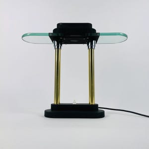 Könnte beinhalten: Eine Vintage-Schreibtischlampe mit schwarzem Sockel und Oberteil, getragen von zwei goldfarbenen Säulen. Die Lampe hat ein klares Glasregal und einen schwarzen oberen Teil. Ein schwarzes Netzkabel geht von der Basis aus.