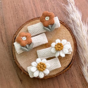 Puede incluir: Cinco pinzas para el pelo hechas a mano con diseños de flores bordadas. Dos pinzas presentan flores naranjas con centros blancos y hojas verdes. Las otras tres pinzas tienen flores blancas con centros amarillos. Las pinzas están unidas a tela blanquecina.