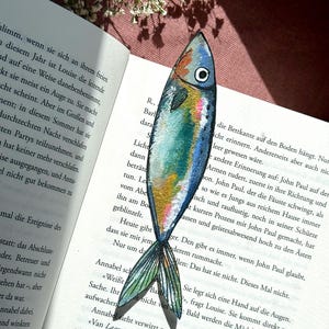 Op de afbeelding: Een handbeschilderde boekenlegger in de vorm van een vis met levendige blauwe, groene en gouden tinten, voorzien van een gedetailleerd oog en vinstructuur. De boekenlegger ligt verticaal op een opengeslagen boekpagina met tekst.