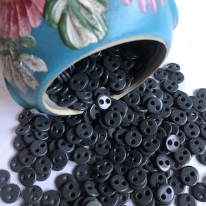 May include: A large bowl filled with dark gray buttons spills out onto a white surface. The buttons are all the same size and shape, with two holes in the center. The bowl is decorated with a floral pattern in shades of pink, blue, and green.