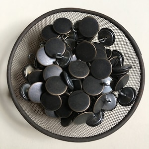 May include: A collection of matte black buttons with a metallic bronze rim, filling a round, dark metal mesh basket. The buttons are circular with a loop on the back for sewing. The basket sits on a light-colored surface.