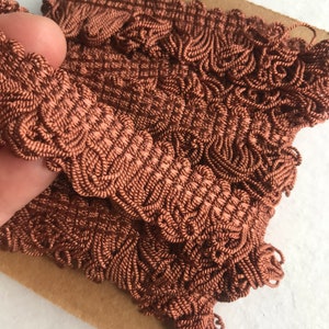May include: Close-up of a roll of brown decorative trim. The trim is made of tightly woven, textured material with a scalloped edge. The color is a warm, reddish-brown, and the trim appears to be new.