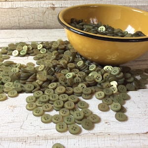 Puede incluir: Un taz&oacute;n grande lleno de botones verdes se derrama sobre una superficie de madera blanca. Los botones son todos del mismo tama&ntilde;o y forma, y tienen cuatro agujeros cada uno.