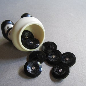 May include: A white ceramic container with a black and white design, tipped over, spilling out black buttons with rhinestones.