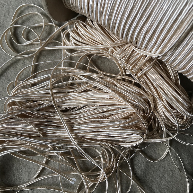 Pode incluir: Um close-up de uma pilha de cord&atilde;o tran&ccedil;ado de cor marfim. O cord&atilde;o &eacute; feito de um material macio e flex&iacute;vel e tem um brilho sutil.