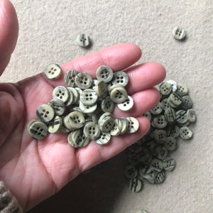 Puede incluir: Una colección de pequeños botones redondos de color beige con vetas negras y blancas. Los botones están esparcidos sobre una mano y una mesa.