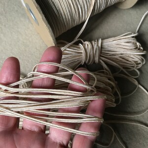 Pode incluir: Um carretel de cord&atilde;o de cetim de cor marfim com um brilho sutil. O cord&atilde;o est&aacute; enrolado em torno de um carretel de cart&atilde;o e est&aacute; sendo segurado em uma m&atilde;o.