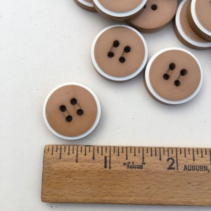 Op de afbeelding: Een groep bruine plastic knoppen met een witte rand. De knoppen hebben elk twee gaten.