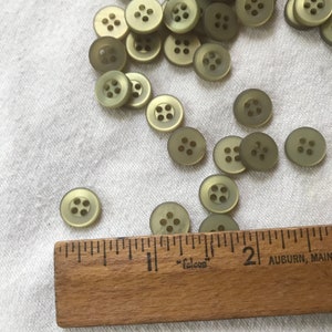 Puede incluir: Primer plano de un grupo de 20 botones de pl&aacute;stico verde con cuatro agujeros cada uno. Los botones est&aacute;n esparcidos sobre una superficie de tela blanca. Una regla de madera con la palabra "falcon" impresa en ella est&aacute; en la parte inferior de la imagen.