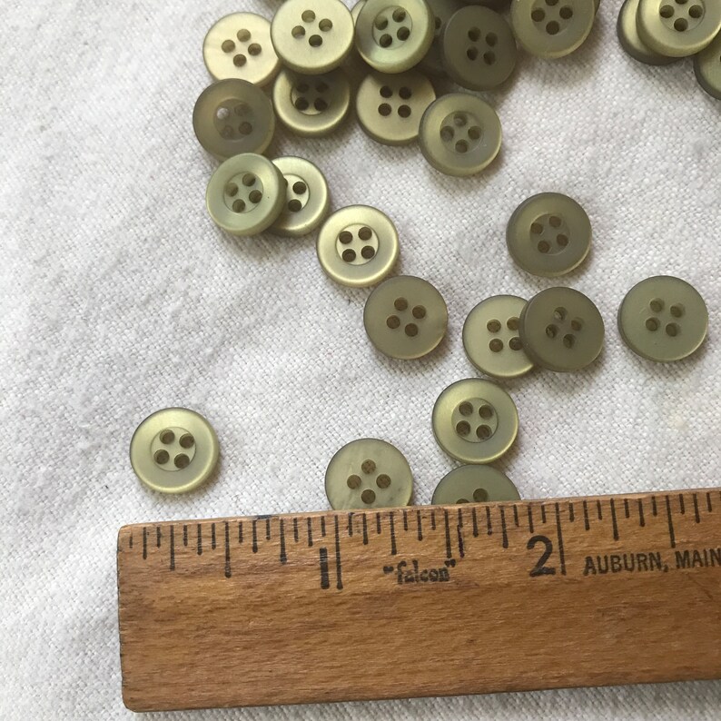 Puede incluir: Primer plano de un grupo de 20 botones de pl&aacute;stico verde con cuatro agujeros cada uno. Los botones est&aacute;n esparcidos sobre una superficie de tela blanca. Una regla de madera con la palabra "falcon" impresa en ella est&aacute; en la parte inferior de la imagen.