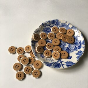 May include: A collection of brown buttons with a white trim, arranged on a blue and white patterned plate. The buttons are all the same size and shape, and they have a two-hole design.