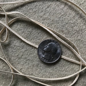 Pode incluir: Um close-up de um cord&atilde;o ou fita branca com um brilho prateado. O cord&atilde;o est&aacute; sobre uma superf&iacute;cie texturizada e envolto em torno de uma moeda de 25 centavos de d&oacute;lar americano, datada de 2021. A moeda de 25 centavos &eacute; usada para mostrar a escala do cord&atilde;o.