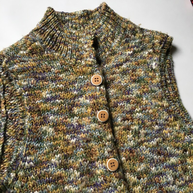 May include: A sleeveless knitted vest with a high neck and a multi-colored striped pattern. The vest has three brown buttons down the front.