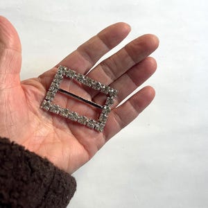 May include: A rectangular silver buckle with a row of clear rhinestones around the edges. The buckle has a silver bar across the center. The buckle is held in a hand.