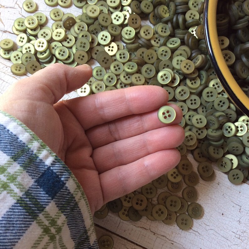 Puede incluir: Un mont&oacute;n grande de botones de pl&aacute;stico verde con cuatro agujeros cada uno. Los botones est&aacute;n esparcidos sobre una superficie blanca. Una mano est&aacute; sosteniendo uno de los botones.