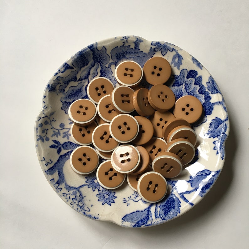 May include: A collection of brown buttons with white trim, arranged in a bowl with a blue and white floral pattern.