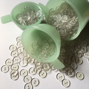 May include: A close-up of a large quantity of clear plastic buttons in various sizes, spilling out of three pale green measuring cups.