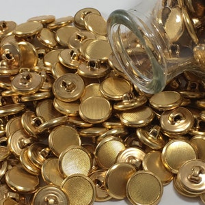 Puede incluir: Un montón grande de botones de metal de color dorado con textura acanalada. Los botones se están derramando de un frasco de vidrio transparente.