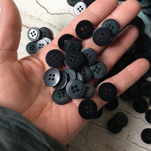 May include: A collection of round, dark charcoal-colored buttons, each featuring four central holes. These circular fasteners are shown in a cluster, displaying a smooth, matte finish and a consistent design suitable for various sewing and craft projects.