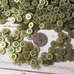 Puede incluir: Un mont&oacute;n grande de botones de pl&aacute;stico verde con cuatro agujeros cada uno. Los botones est&aacute;n esparcidos sobre una superficie blanca.