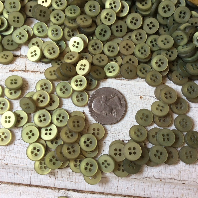 Puede incluir: Un mont&oacute;n grande de botones de pl&aacute;stico verde con cuatro agujeros cada uno. Los botones est&aacute;n esparcidos sobre una superficie blanca.