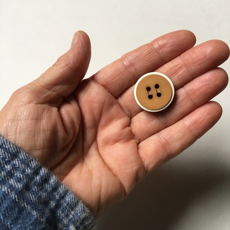 May include: A brown button with two holes, surrounded by a white ring. The button is being held in a hand.
