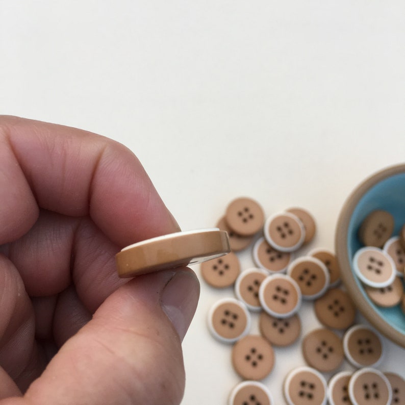 Op de afbeelding: Een close-up van een bruine knoop met een witte rand. De knoop heeft vier gaten en is omringd door andere vergelijkbare knoppen.