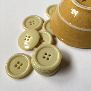 May include: A group of off-white plastic buttons with four holes each. The buttons are arranged in a bowl, with some spilling out onto a white surface.